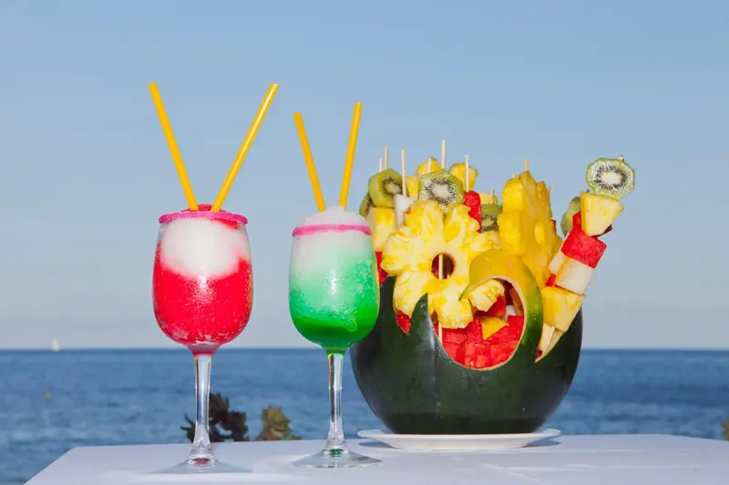Deux cocktails colorés et une présentation de fruits exotiques sur une table ensoleillée.