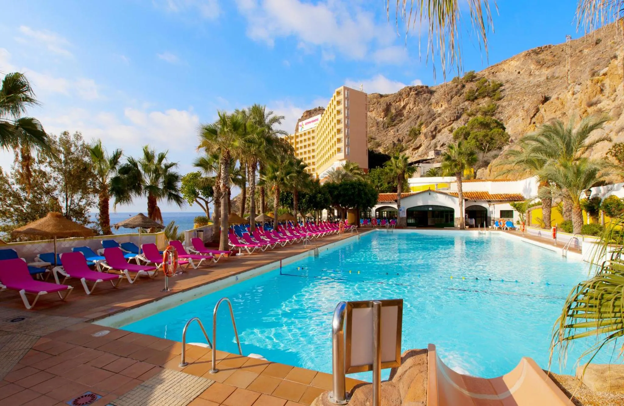 Une piscine turquoise bordée de transats roses, avec un bâtiment et des palmiers en arrière-plan.