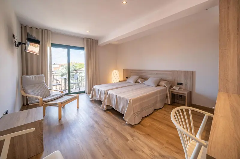 Chambre lumineuse avec un lit double et un fauteuil près d'une baie vitrée.