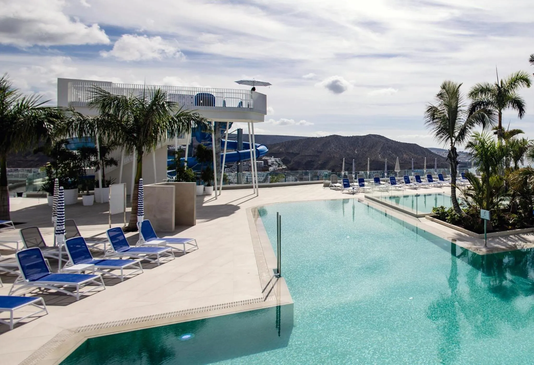 Une piscine rectangulaire bordée de transats blancs et bleus offre une vue sur la mer et les montagnes.