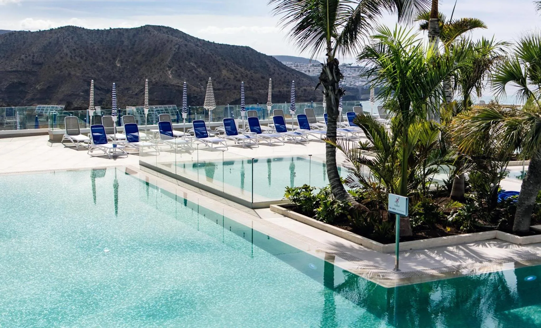 Transats bleus alignés au bord d'une piscine turquoise avec vue sur la mer et les montagnes.