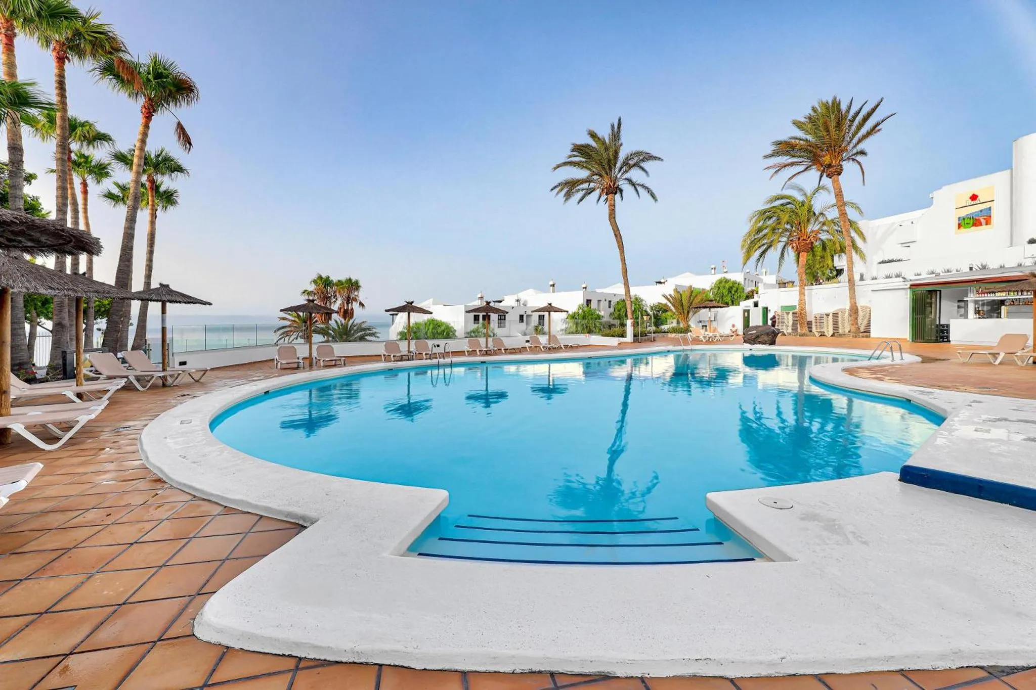 Piscine rectangulaire entourée d'un sol carrelé et de transats, avec des palmiers en arrière-plan.