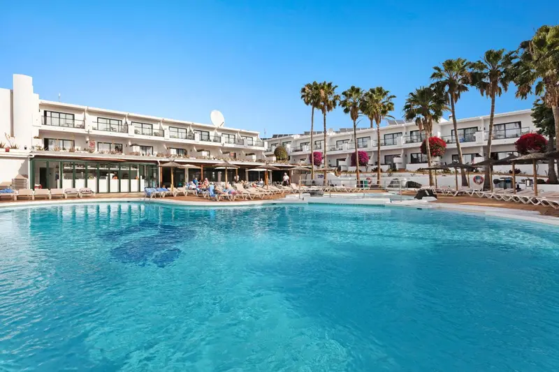 Piscine turquoise devant un bâtiment blanc bordé de palmiers sous un ciel bleu.