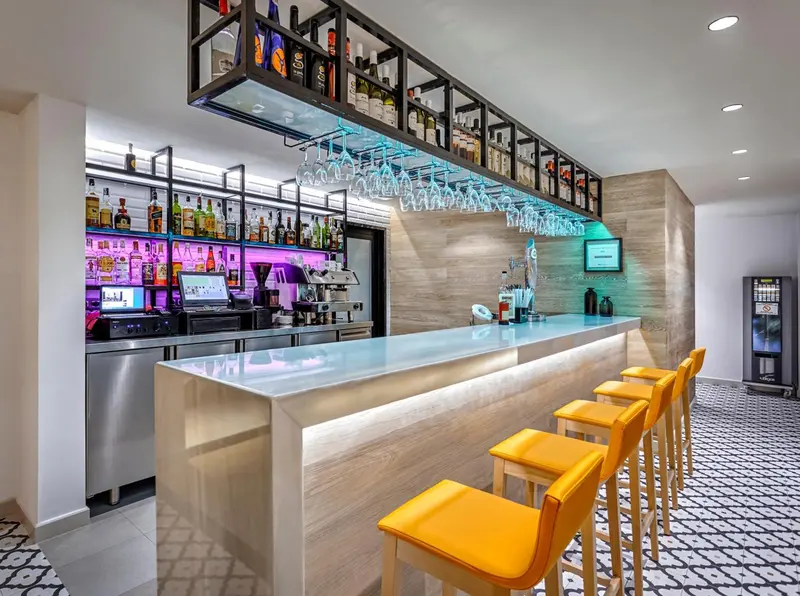 Un comptoir de bar moderne avec des tabourets jaunes et une étagère illuminée de bouteilles.