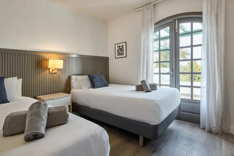 Une chambre lumineuse avec deux lits simples et une porte-fenêtre ouverte sur l'extérieur.