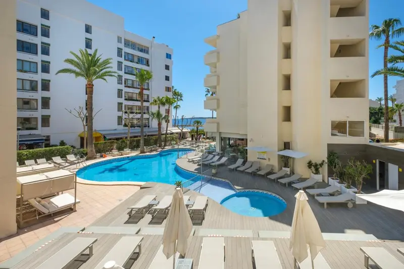 Vue aérienne d'une piscine d'hôtel avec des transats et des parasols, entourée d'une végétation luxuriante et de bâtiments blancs.