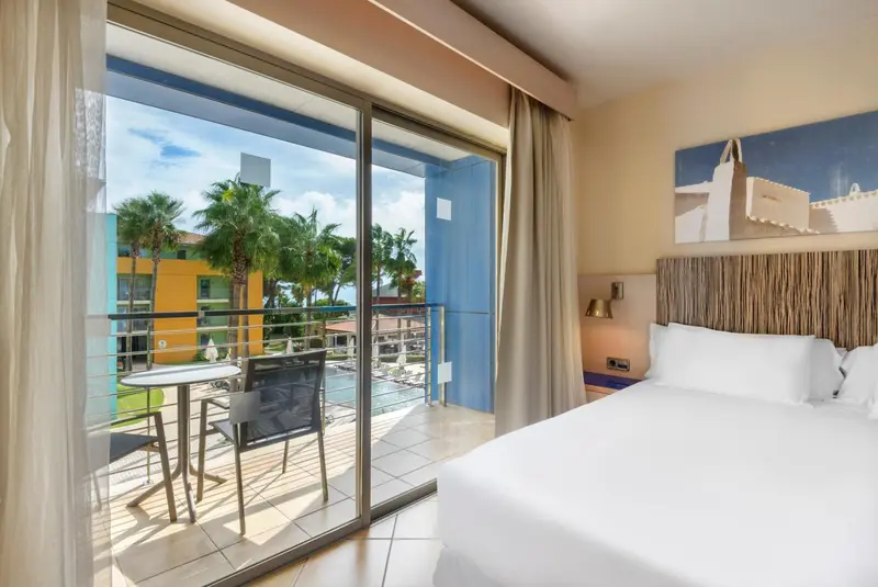 Une chambre lumineuse s'ouvre sur une terrasse avec vue sur la végétation et une piscine.