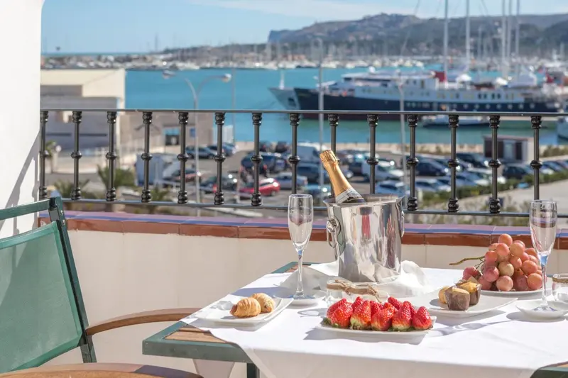 Une table avec des fruits, des viennoiseries et des boissons sur une terrasse avec vue sur un port de plaisance.