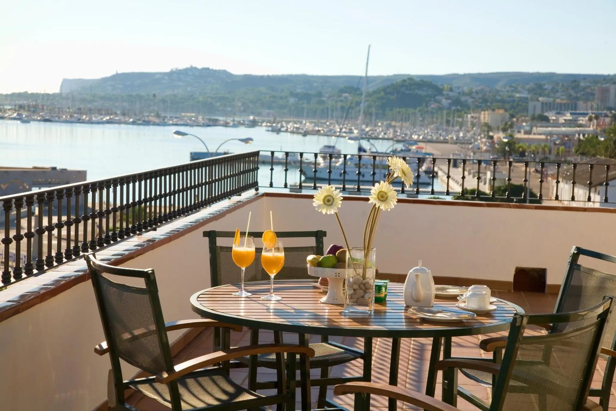 Une table ronde avec des boissons et des fruits est mise en scène sur une terrasse ensoleillée avec vue sur la mer.