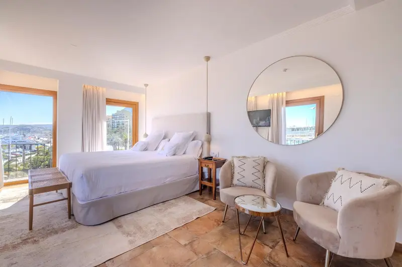 Chambre lumineuse avec un grand lit et un coin salon près d'une fenêtre.