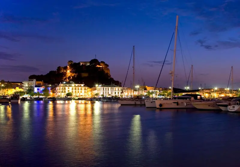 Vue nocturne d'un port de plaisance avec des bateaux à quai et une colline illuminée en arrière-plan.