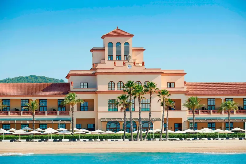 Bâtiment beige à plusieurs étages avec des palmiers et une plage ensoleillée.