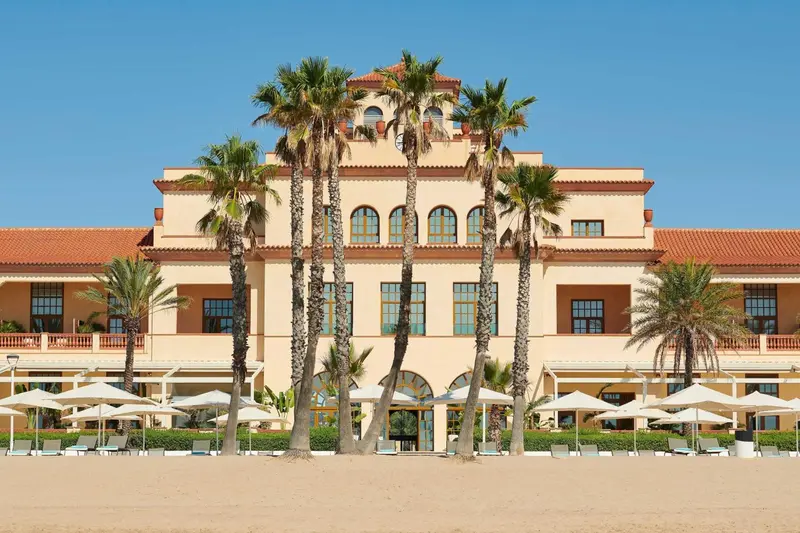 Façade d'un bâtiment beige avec des palmiers et des parasols sur une plage de sable.