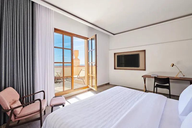Une chambre lumineuse avec un lit blanc et un accès à une terrasse ensoleillée.