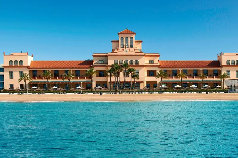Bâtiment beige avec toits en tuiles rouges face à une plage de sable fin et à une mer turquoise.