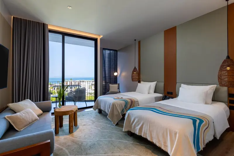 Chambre lumineuse avec deux lits et vue sur la mer par de grandes baies vitrées.