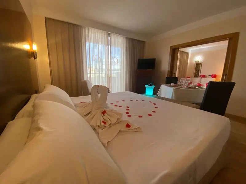 Une chambre d'hotel avec un lit decoré de serviettes en forme de cygnes et une table preparée pour un repas.