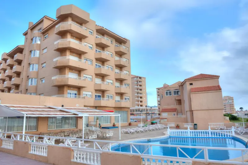Bâtiment beige à plusieurs étages avec des balcons et une piscine en contrebas.