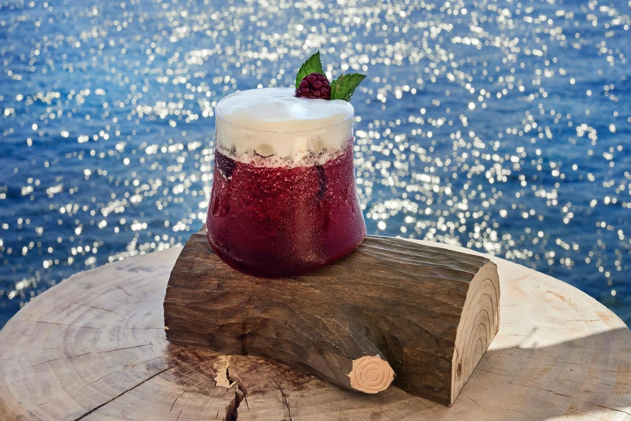 Un cocktail aux fruits rouges est présenté sur une tranche de bois, avec la mer scintillante en arrière-plan.