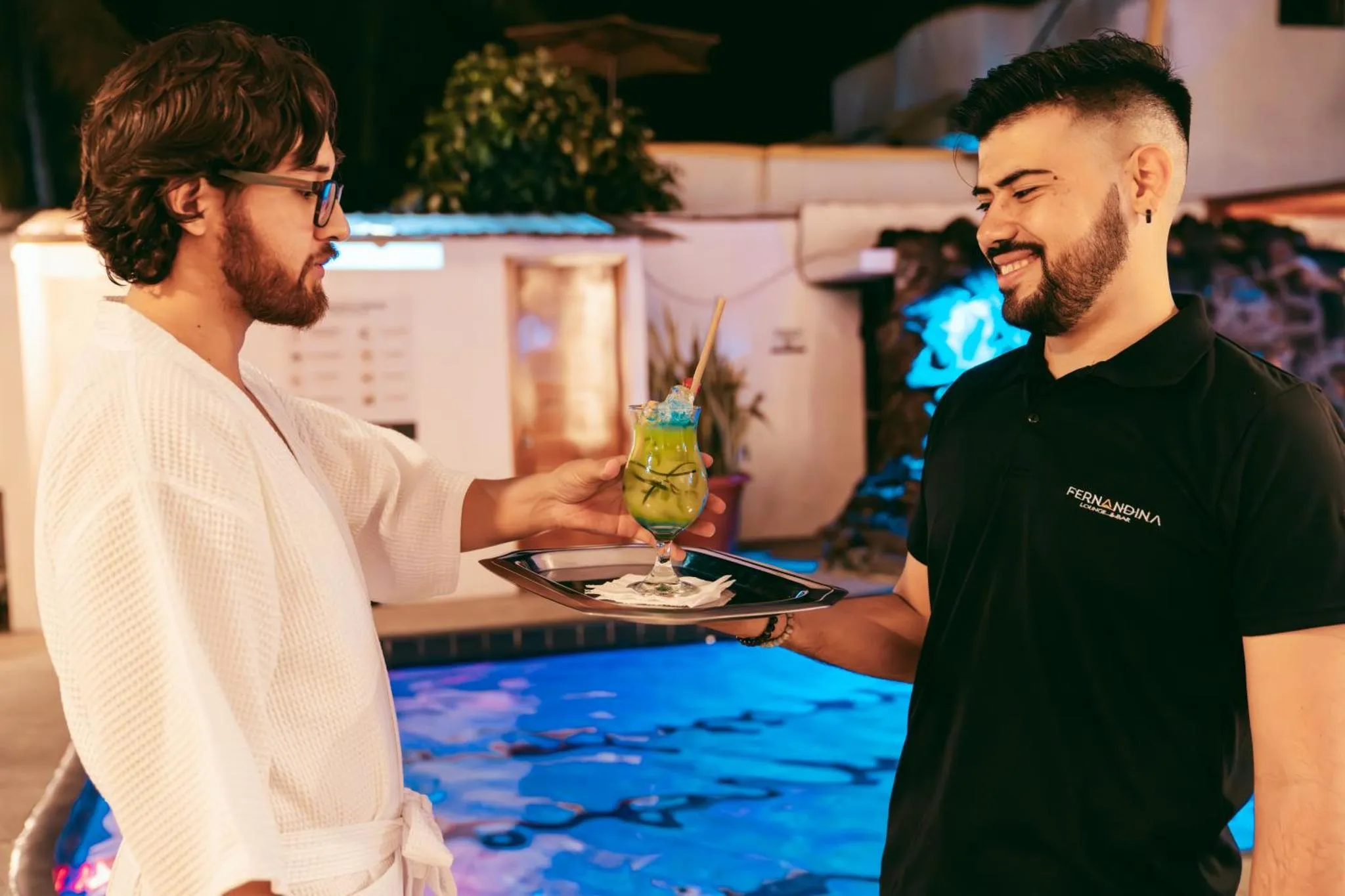 Deux hommes discutent près d'une piscine illuminée, l'un servant un cocktail à l'autre.