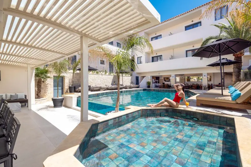 Une femme se relaxe au bord d'une piscine ensoleillée, entourée de chaises longues et de végétation.