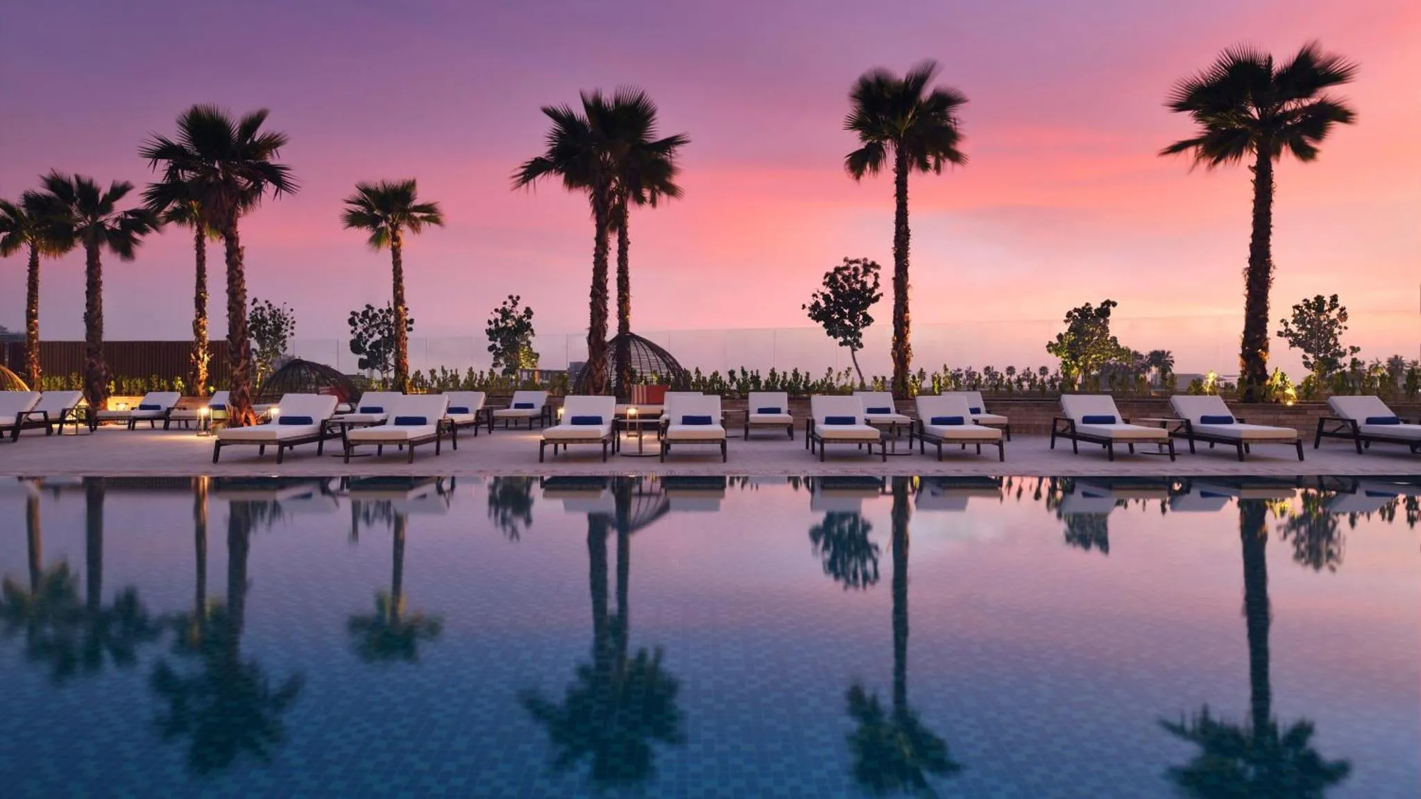 Transats alignés au bord d'une piscine reflétant des palmiers sous un ciel rose au coucher du soleil.