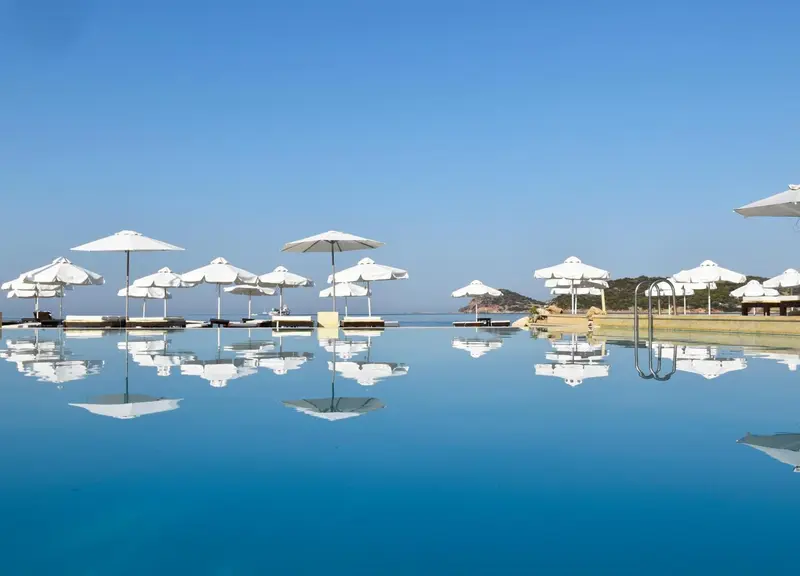 Une piscine à débordement reflète le ciel bleu et les parasols blancs.