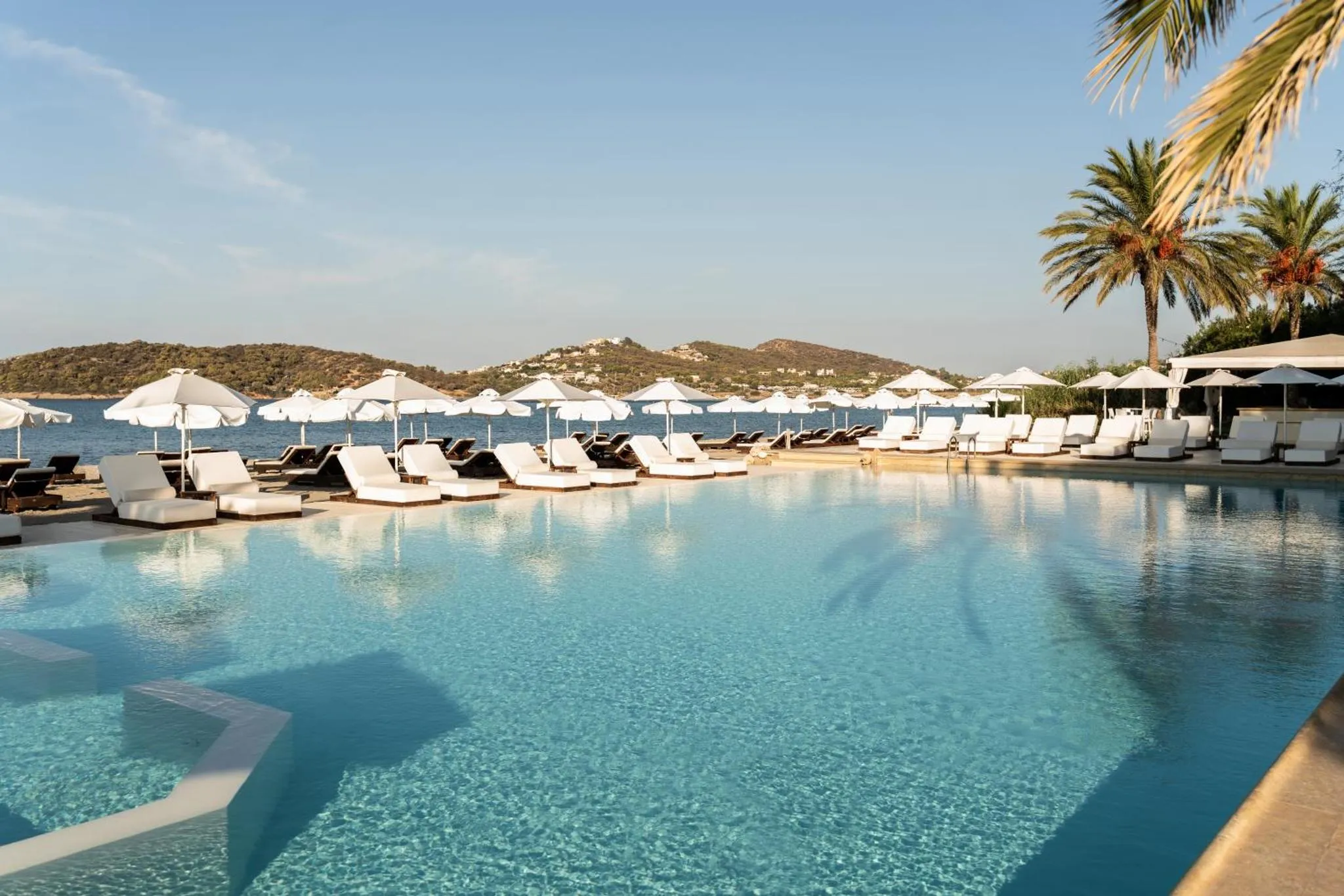 Transats blancs alignés au bord d'une piscine turquoise, avec des parasols et une vue sur la mer.