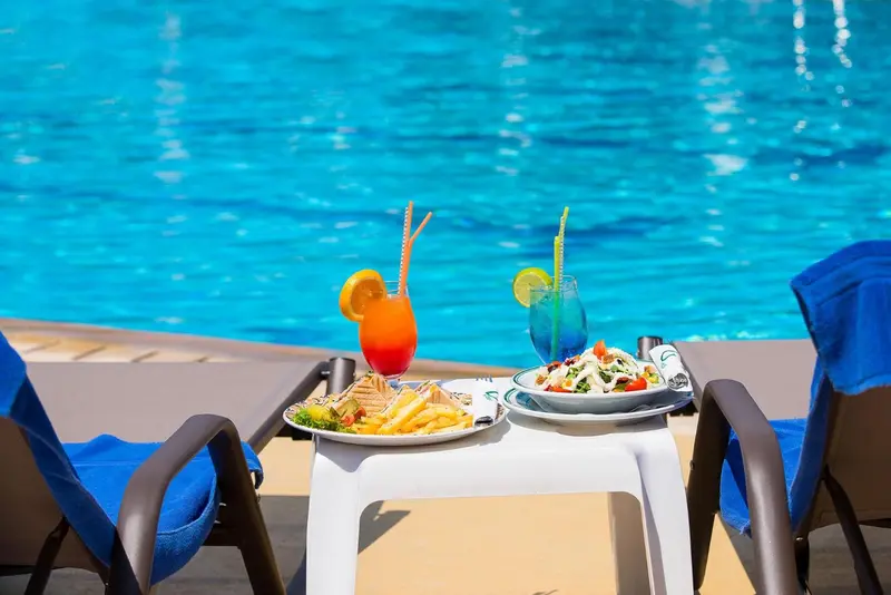 Deux cocktails colorés et des assiettes de nourriture sont servis sur une table au bord d'une piscine.
