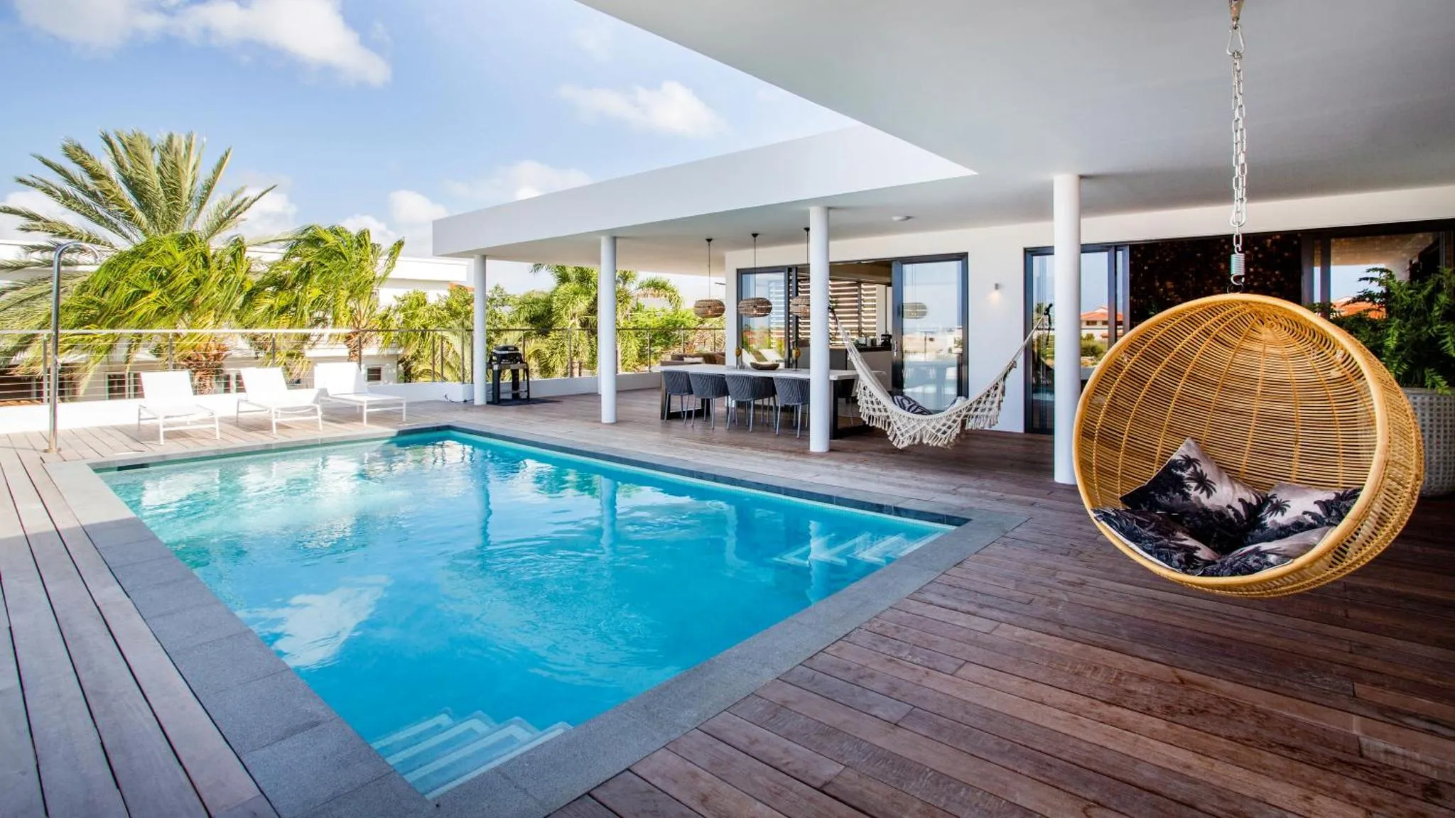 Une piscine rectangulaire bordée d'une terrasse en bois avec des chaises longues et un hamac suspendu.