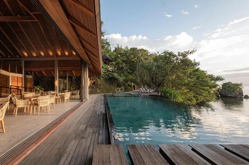 Terrasse en bois avec tables et chaises donnant sur une piscine et la mer.