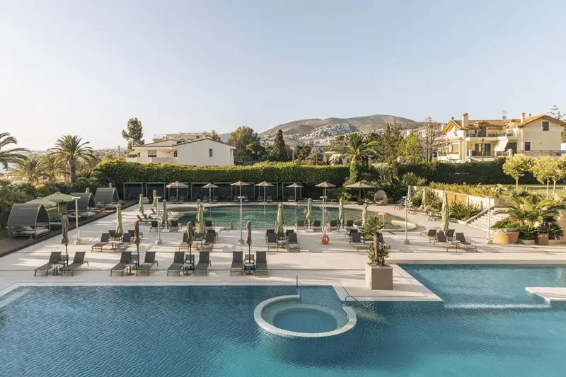 Vue aérienne d'une piscine avec des transats et des parasols, entourée d'une végétation luxuriante et de bâtiments.