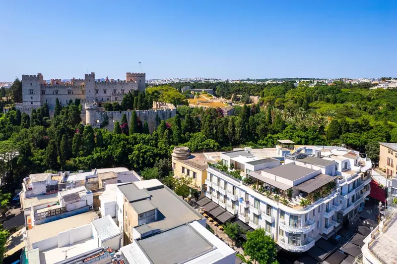 Vue aérienne d'un bâtiment blanc moderne et d'un château médiéval entourés de végétation luxuriante.