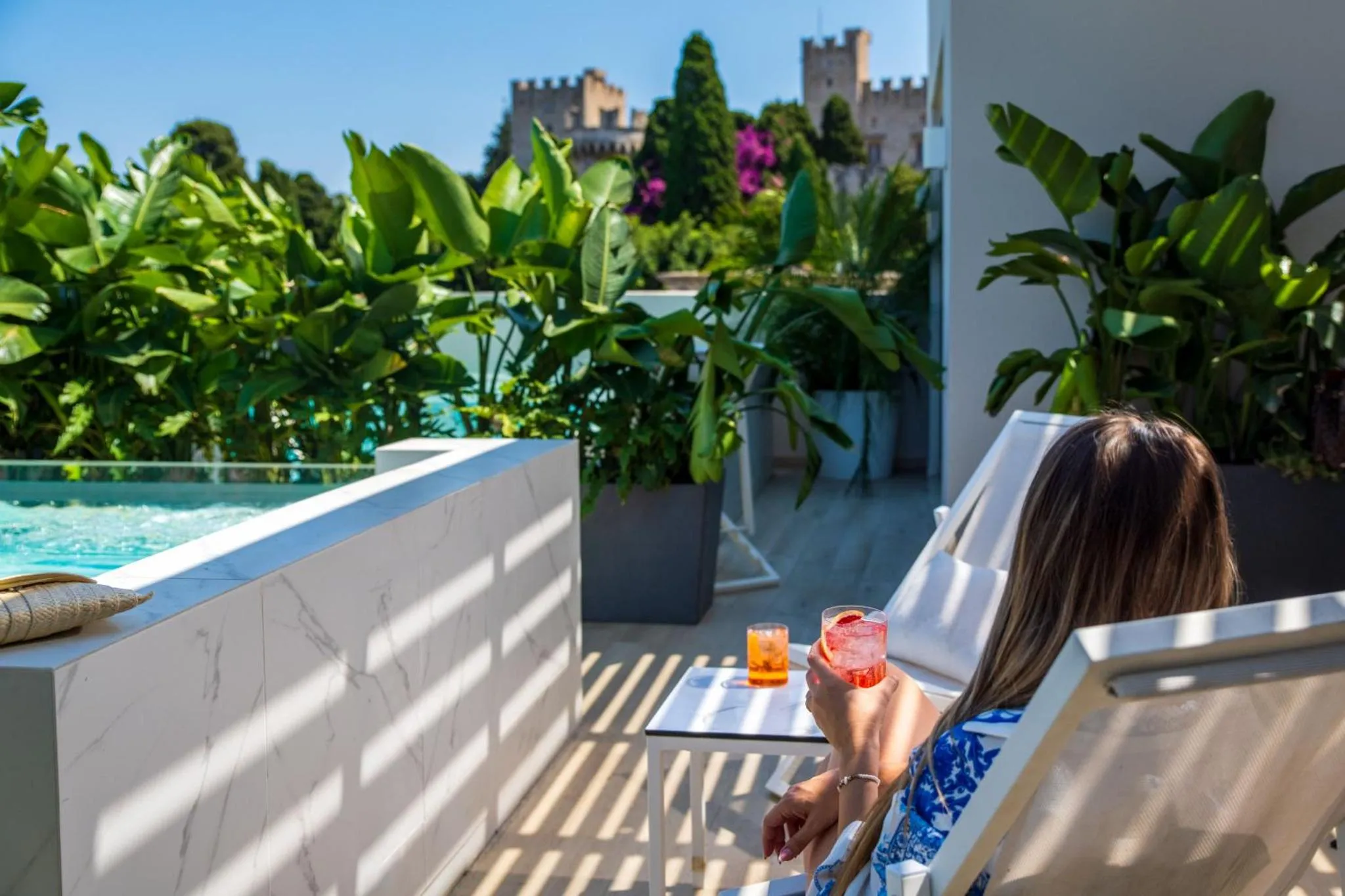 Une personne se détend sur une chaise longue avec un verre, près d'une piscine et de plantes luxuriantes.