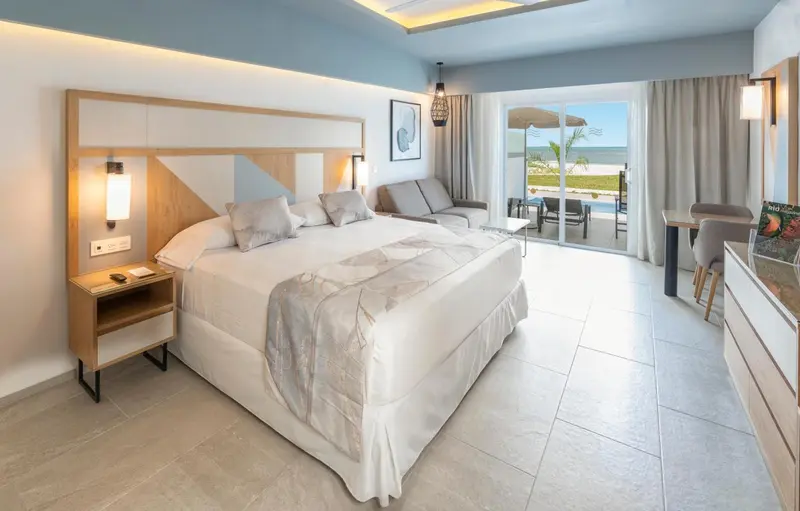 Une chambre lumineuse avec un grand lit et un coin salon donnant sur une piscine.