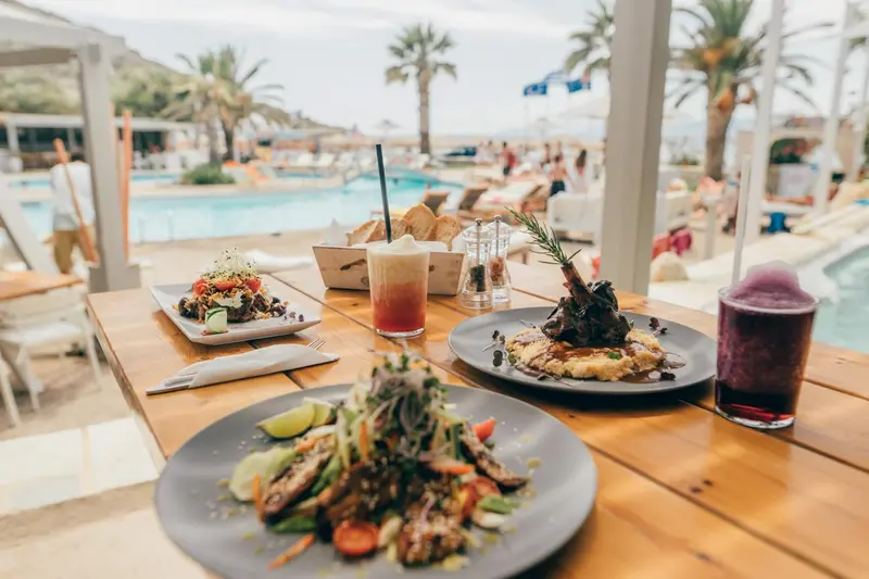 Une table en bois présente des plats colorés et des boissons rafraîchissantes en bord de piscine.