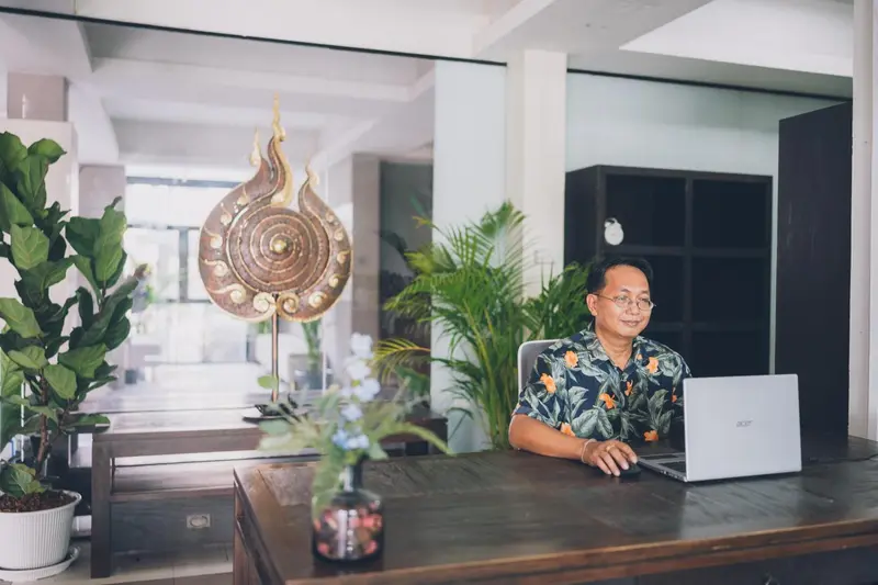 Un homme est assis à un bureau avec un ordinateur portable, devant une décoration florale et une statue.