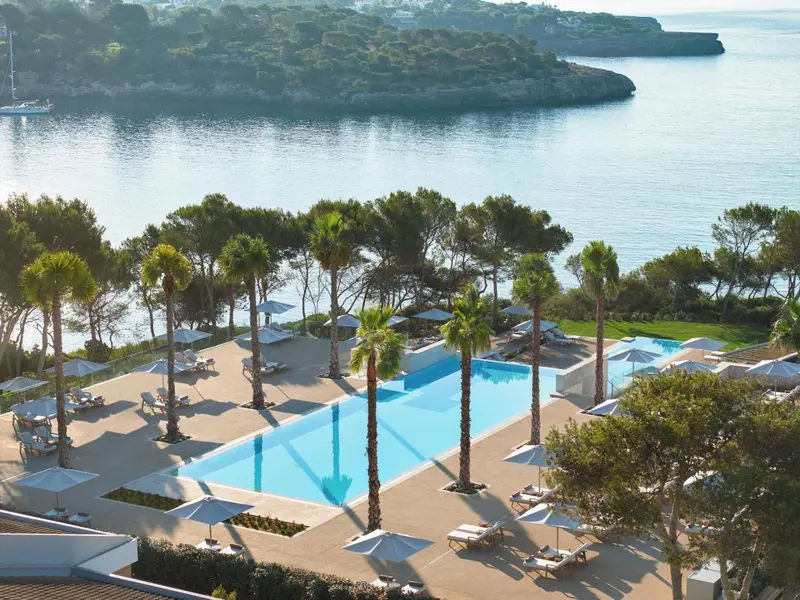 Vue aérienne d'une piscine d'hôtel bordée de palmiers et de transats, surplombant une crique.