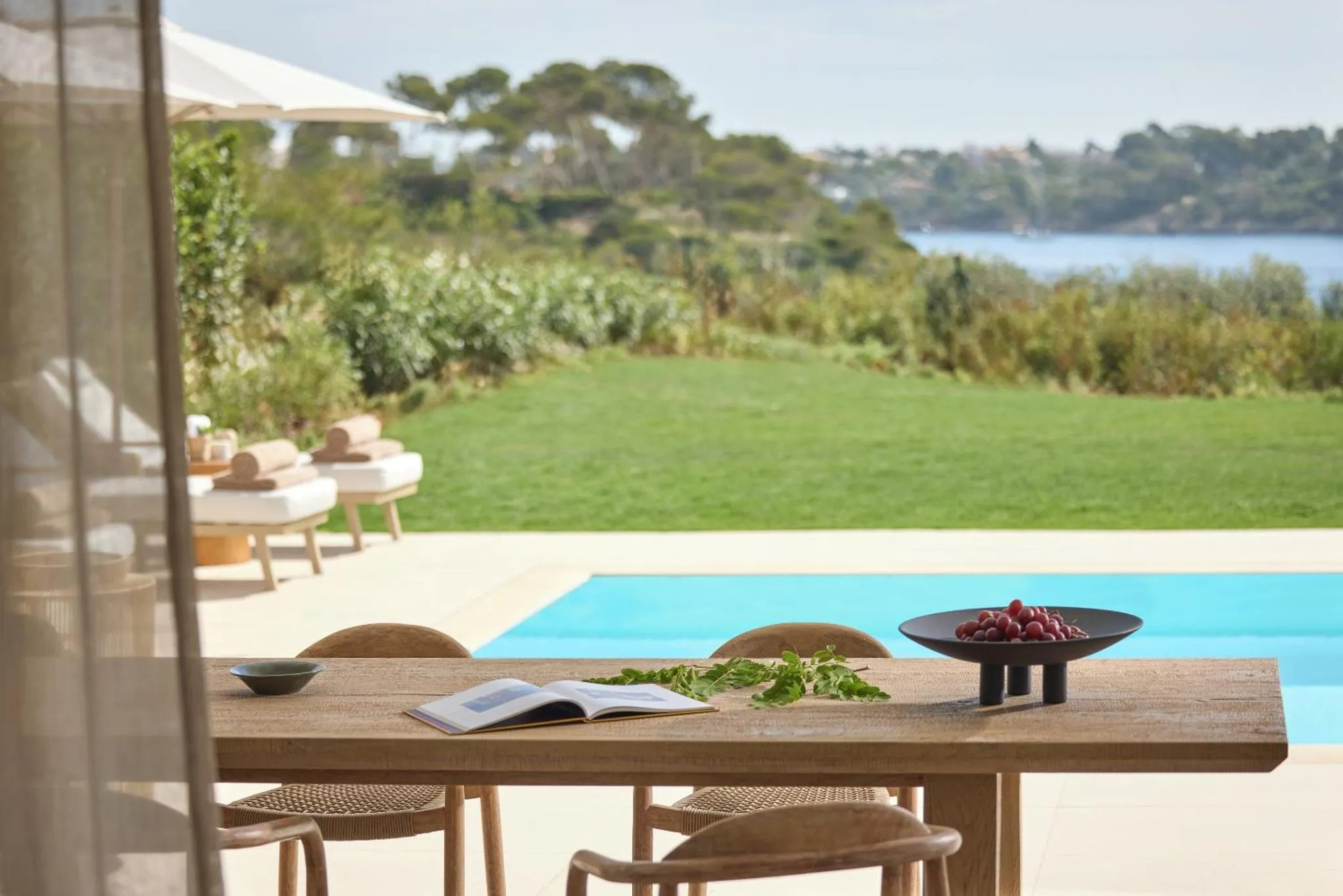Table de repas en bois avec un livre ouvert et des fruits frais, devant une piscine et une vue sur la mer.