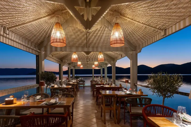 Une salle à manger extérieure avec des tables dressées face à la mer au coucher du soleil.