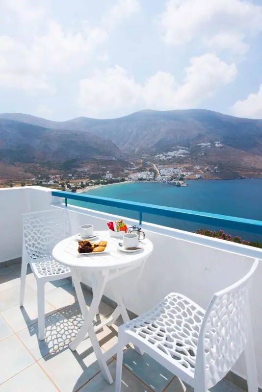 Une table ronde blanche avec des couverts est posée sur une terrasse ensoleillée avec vue sur la mer et les montagnes.