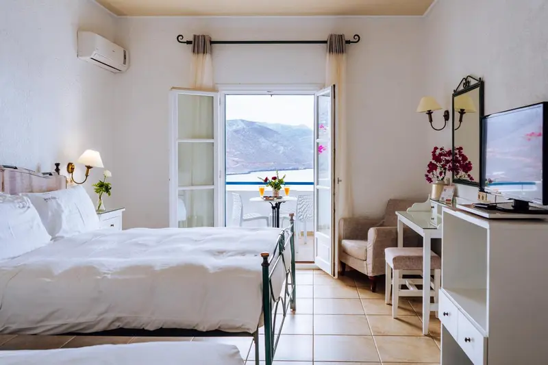 Chambre lumineuse avec un lit blanc et une vue sur la mer depuis un balcon.