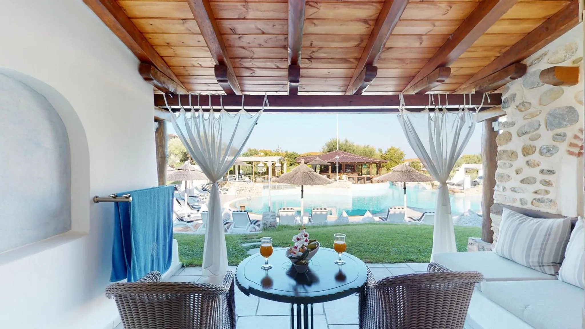 Une table ronde avec des boissons est mise en scène sur une terrasse ombragée donnant sur une piscine et un jardin.