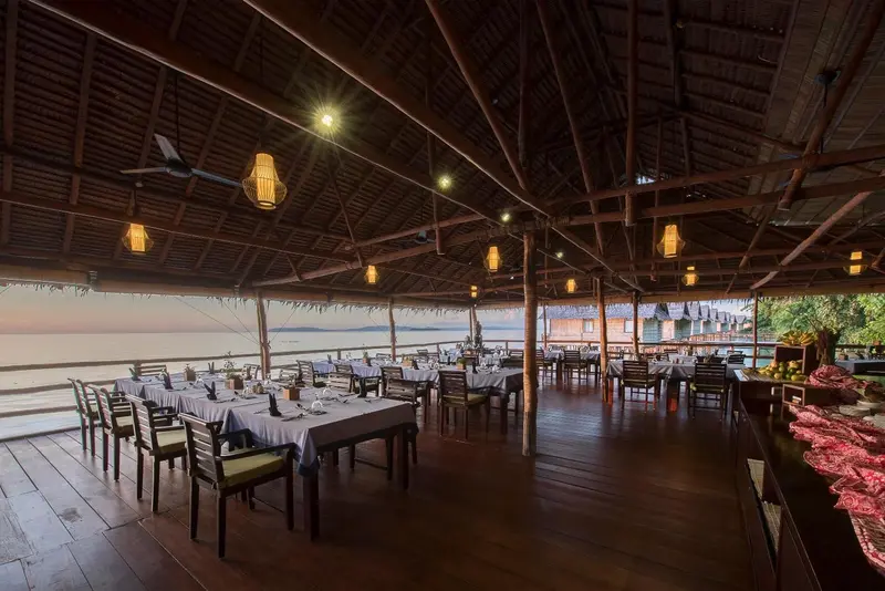 Une salle à manger ouverte sur la mer avec des tables dressées et un éclairage chaleureux.