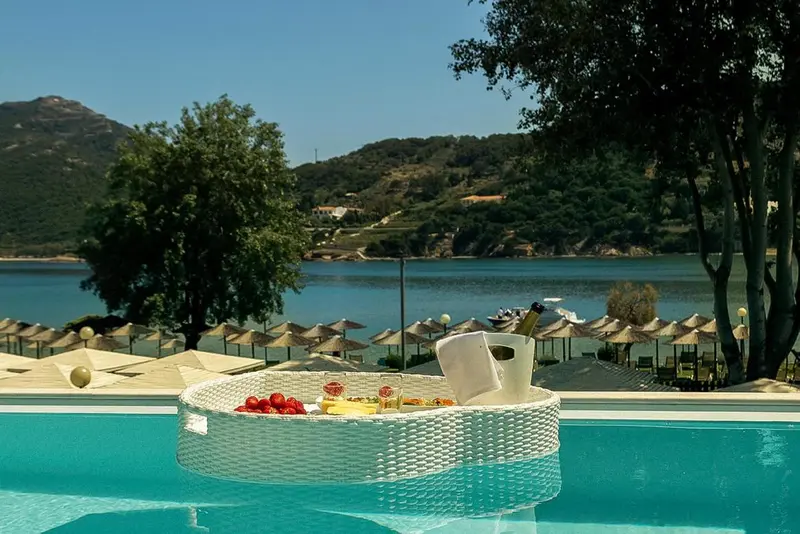 Une table basse en osier flottant dans une piscine turquoise offre une sélection de fruits et une bouteille de champagne.