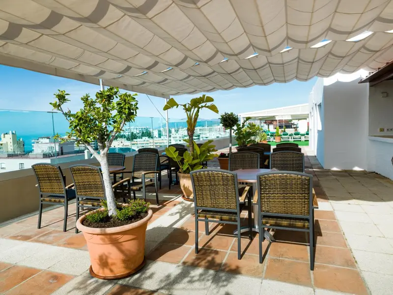 Terrasse ombragée avec tables et chaises donnant sur une vue urbaine et la mer.