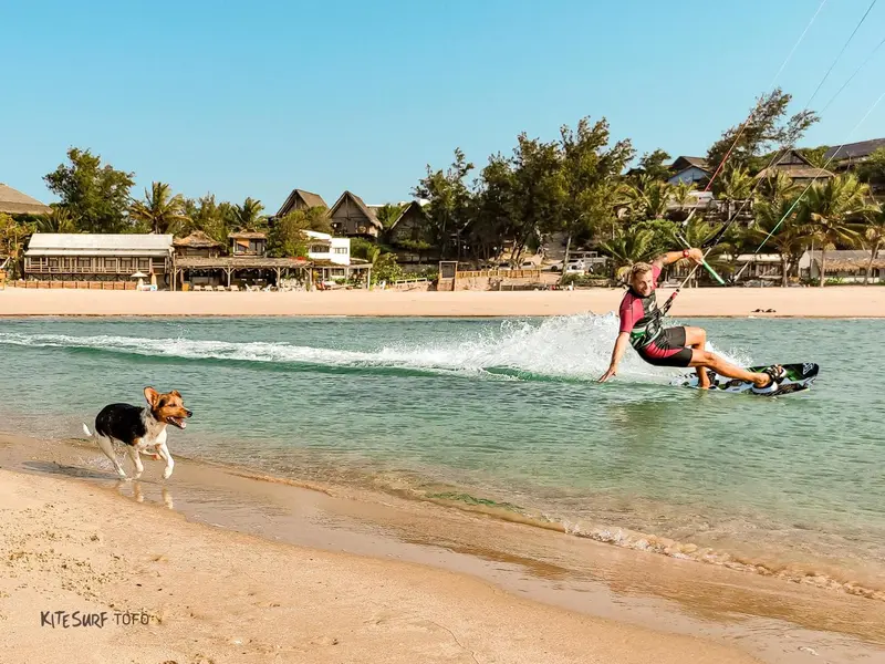 Un kitesurfeur s'envole au-dessus de l'eau turquoise, poursuivi par un chien sur la plage.