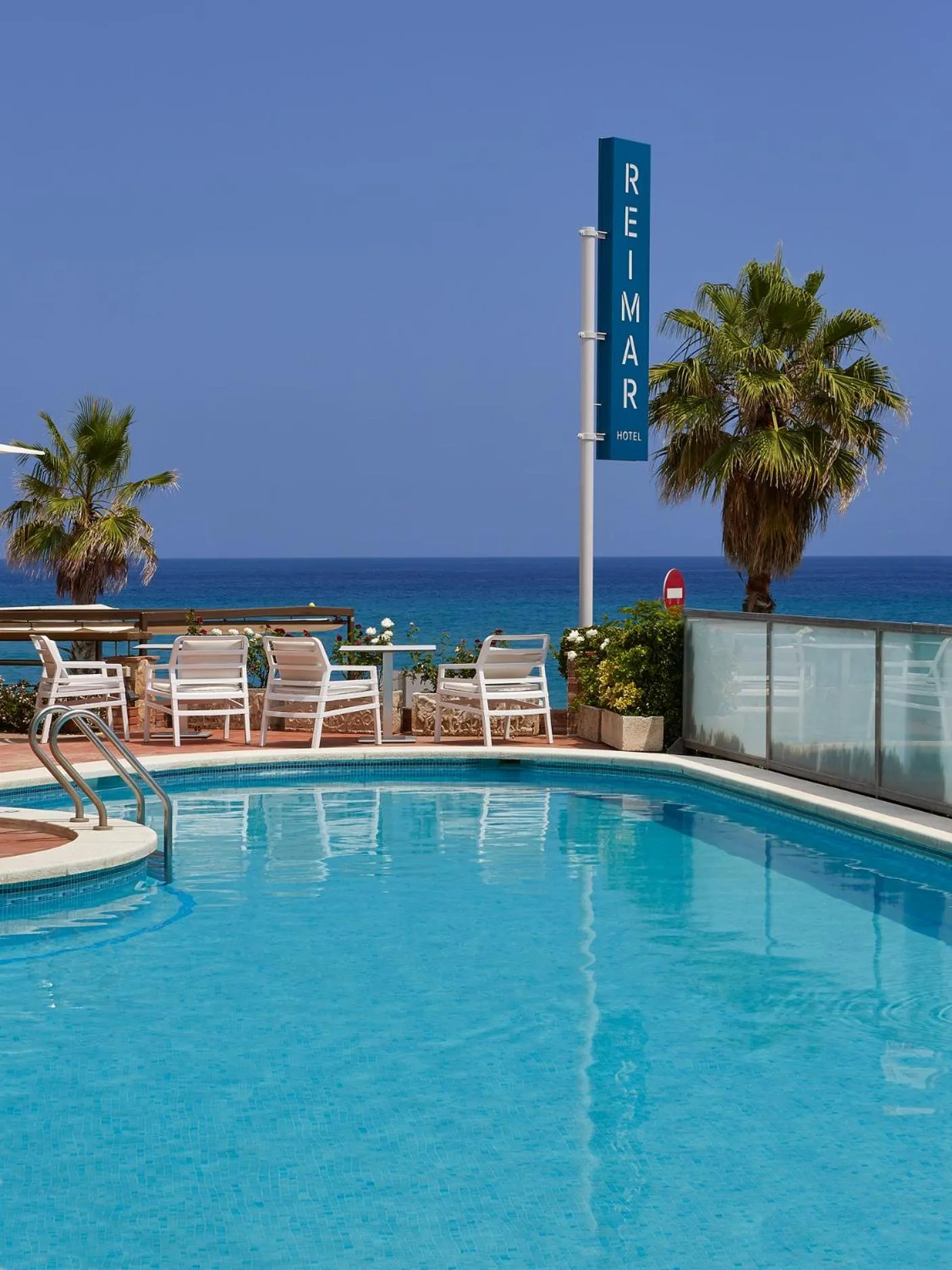 Une piscine turquoise bordée de chaises longues et de palmiers, avec une vue sur la mer.