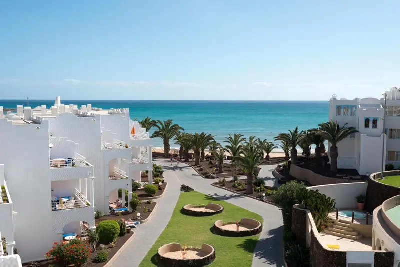 Vue aérienne d'un complexe hôtelier blanc avec piscine et palmiers face à la mer.