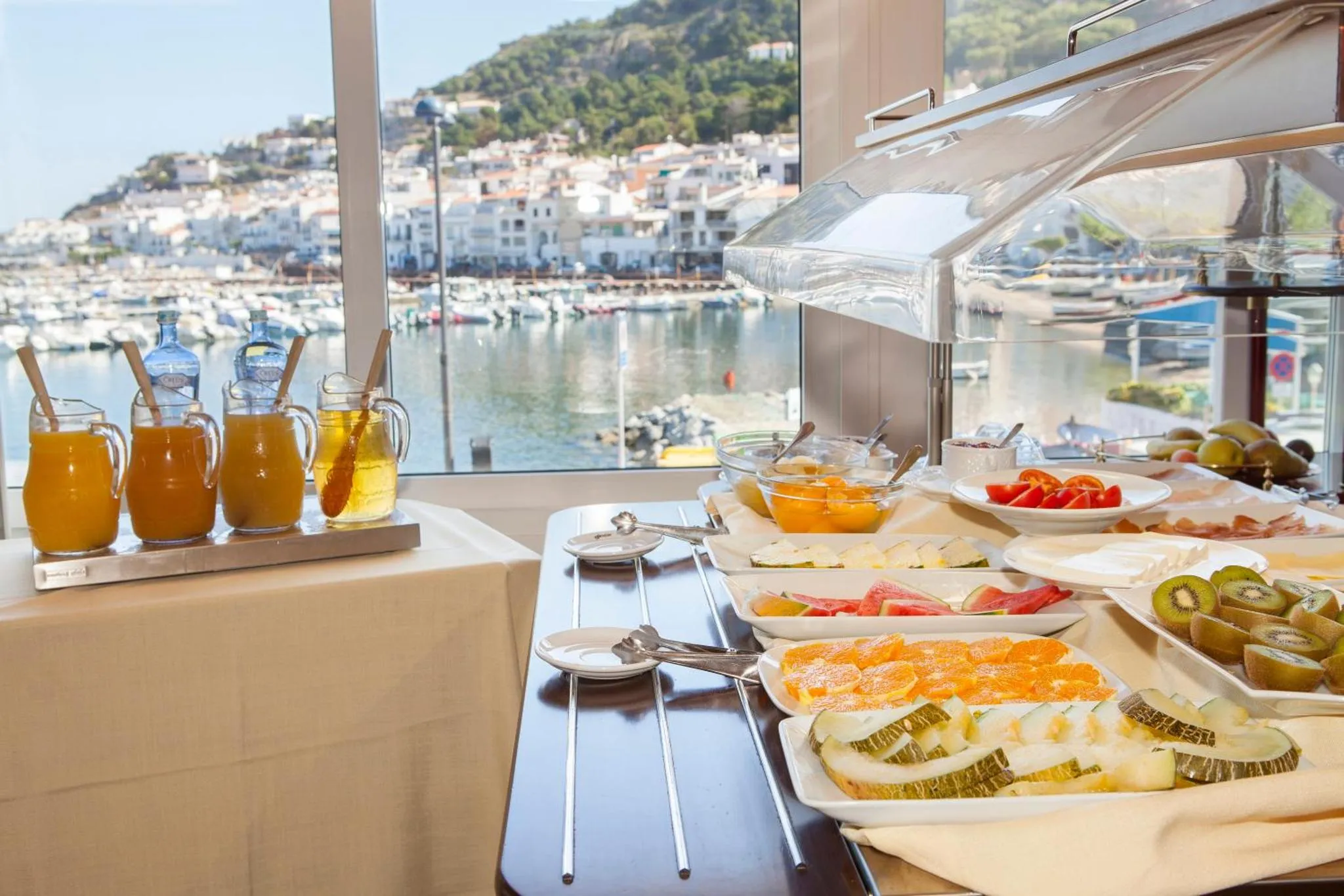 Un buffet propose des jus de fruits, des pâtisseries et des fruits frais devant une vue sur un port de plaisance.